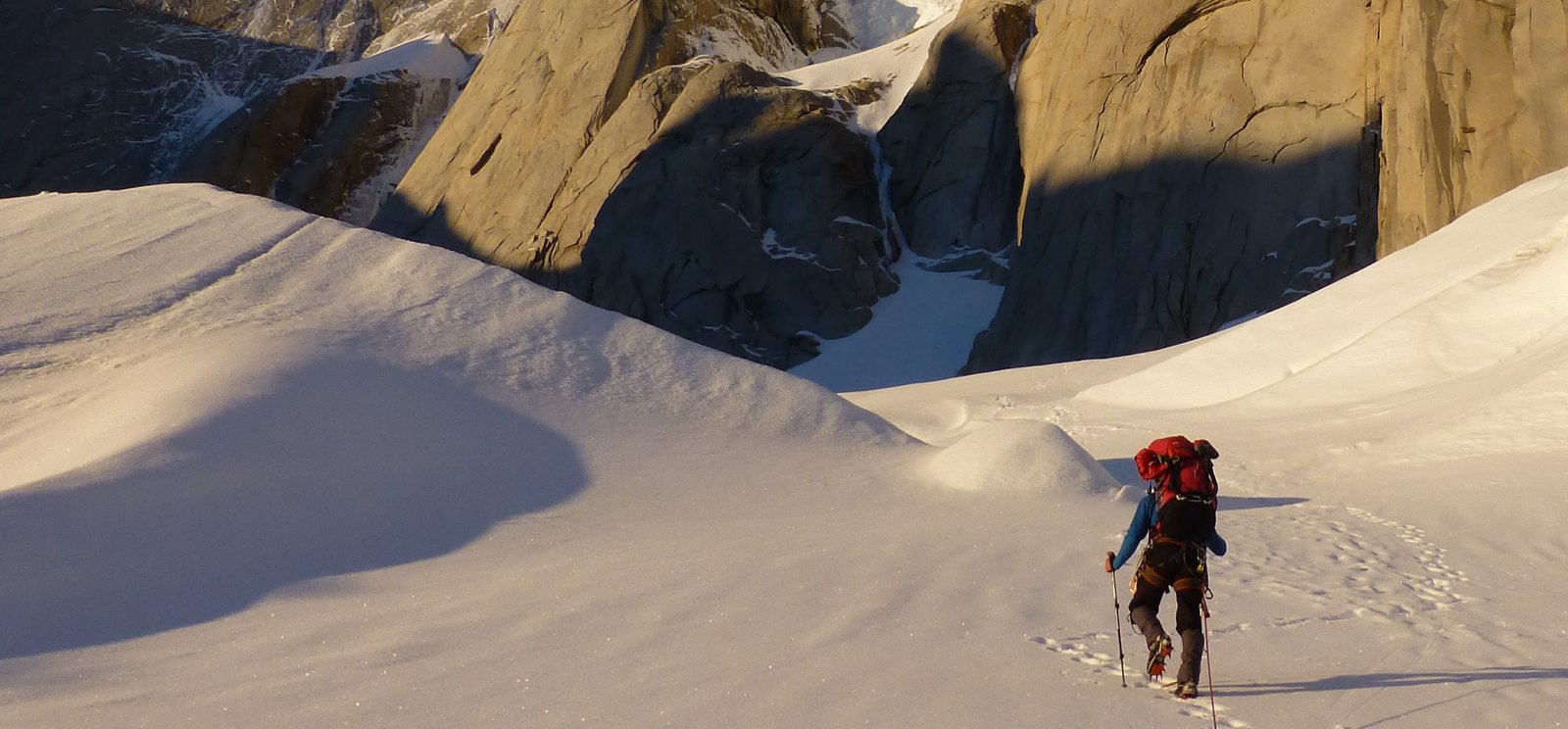 A mountaineer carrying a CRUX pack across snow beneath granite cliffs